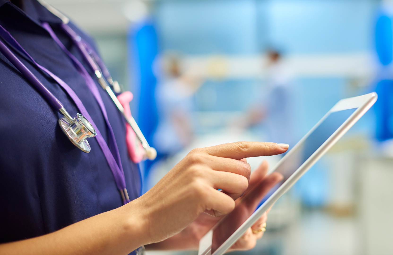 A UK nurse holds a digital tablet in a busy hospital ward